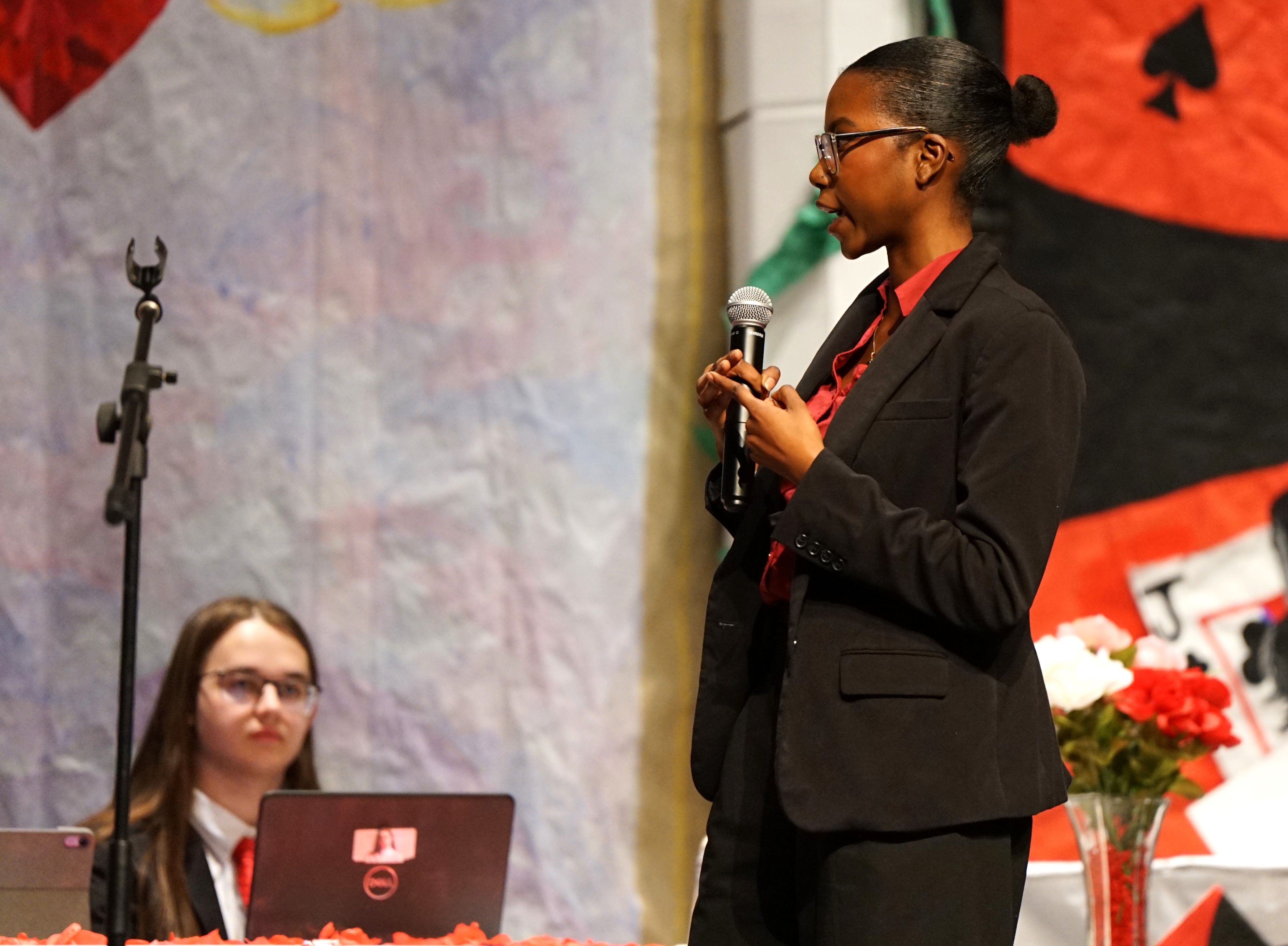 A girl in a black suit holds a microphone as she debates on stage