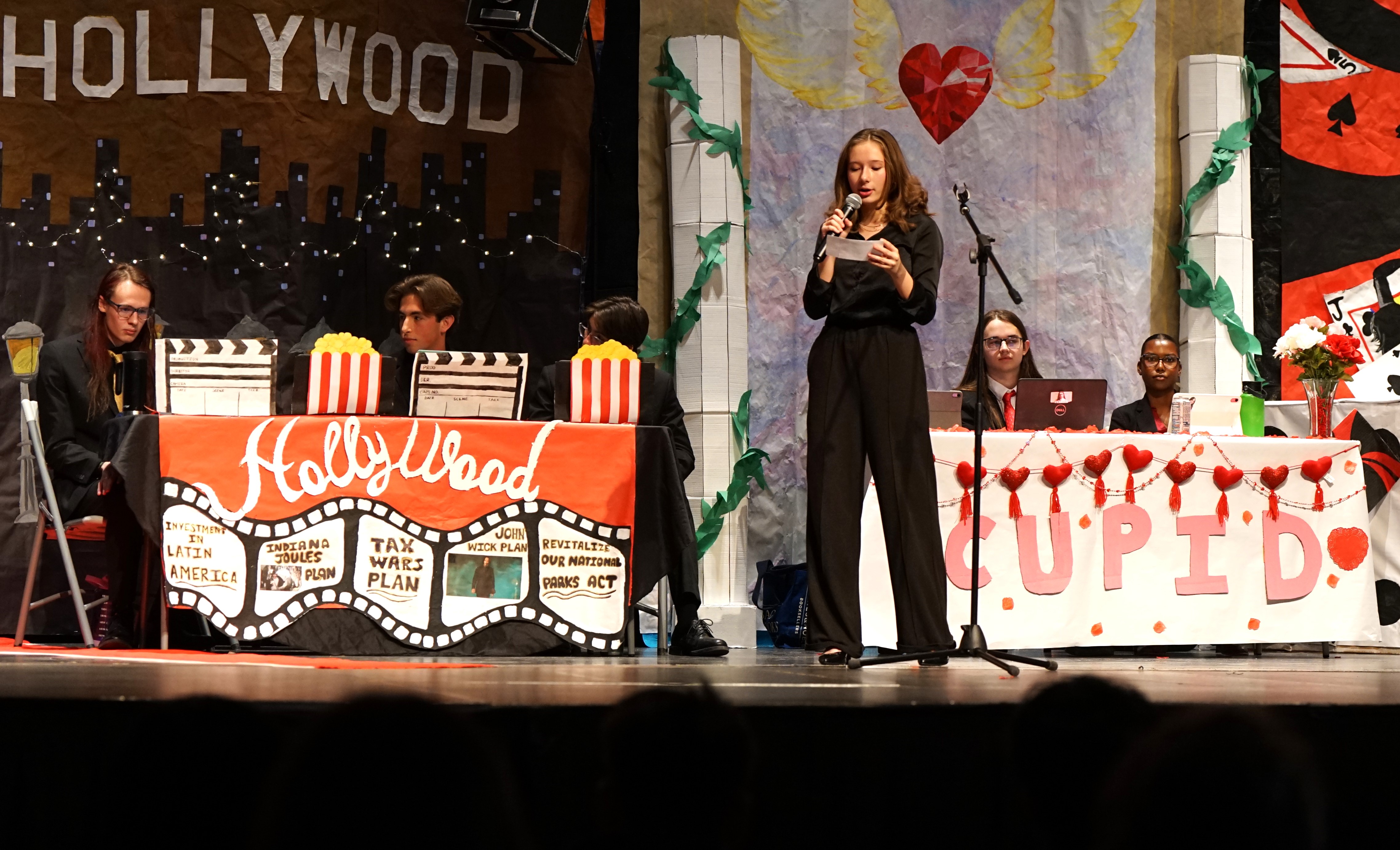 A girl in a black suit stands in front of tables of other students during the debate