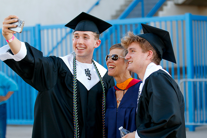 Two teen boys in a black cap and gown take a selfie with a woman in the middle