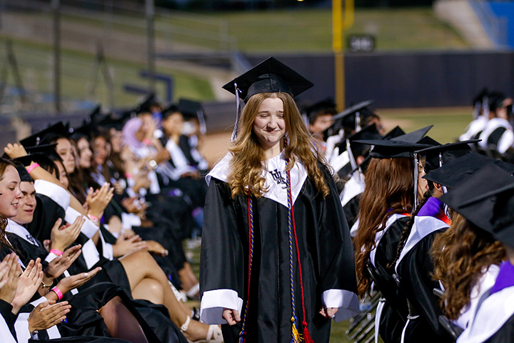 A teen girl in a black cap and gown smiles as she's cheered on to the stage by her fellow graduates