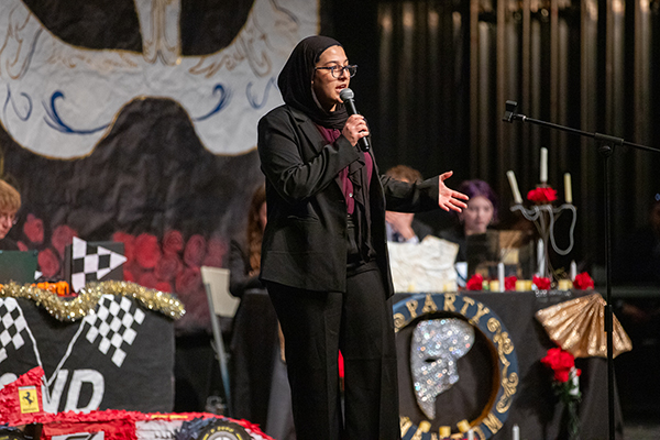 A teen girl in glasses and a black head scarf speaks into a microphone on stage