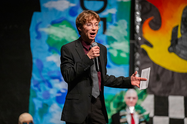 A teen boy in glasses and a black suit speaks into a microphone on stage