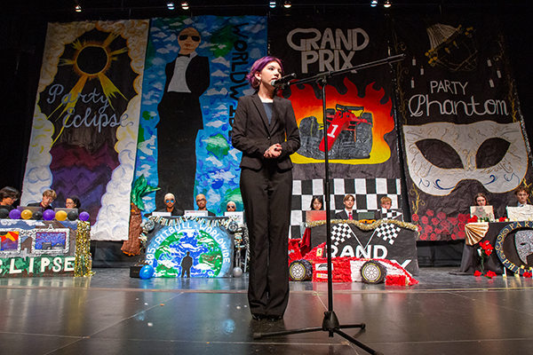A teen girl with short purple hair wearing a black pantsuit speaks into a microphone on stage