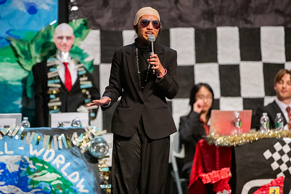 A teen girl wearing a skull cap and sunglasses speaks into a microphone on stage